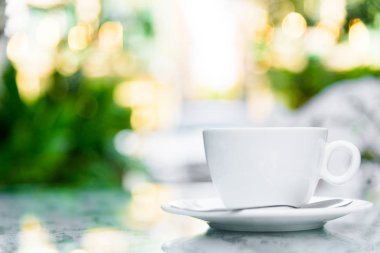 Sabah servis edilecek beyaz bir fincan kahve ve gümüş kaşık. Rahatlatıcı ve sarı ışıklandırma bokeh arka planlı..