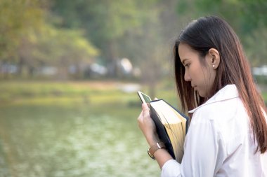 Güzel Asyalı kadın açık havada akıllı telefon kullanıyor ve akşamları elinde günbatımında bahçe parkında kitap tutuyor..