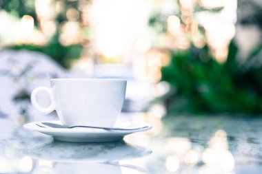 Sabah servis edilecek beyaz bir fincan kahve ve gümüş kaşık. Rahatlatıcı ve sarı ışıklandırma bokeh arka planlı..