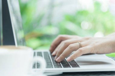Asyalı kadın elleri ve evlilik yüzüğü laptopta kahve, bilgisayar ve pencerenin dışındaki manzarayla dokunup daktilo ediyor..