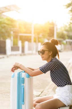 Güzel gezgin Asyalı kadın, büyük beyaz ve mavi bir çantayla sokakta oturuyor. Akşam vakti taksi şoförü bekliyor. Gün batımında..