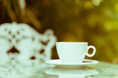 Sabah servis edilecek beyaz bir fincan kahve ve gümüş kaşık. Rahatlatıcı ve sarı ışıklandırma bokeh arka planlı..
