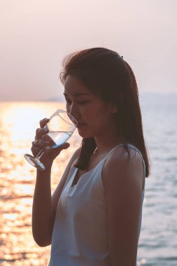 Güzel Asyalı kadın gün batımı ve silüeti ile akşam zamanında deniz ve plajda bir bardak şarap ve içme tutuyor. rahatlatıcı, moda, yaşam tarzı, tatil ve tatil zamanı kavramı.