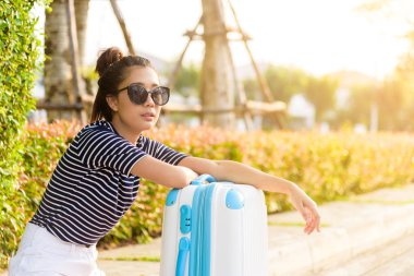 Güzel gezgin Asyalı kadın, büyük beyaz ve mavi bir çantayla sokakta oturuyor. Akşam vakti taksi şoförü bekliyor. Gün batımında..