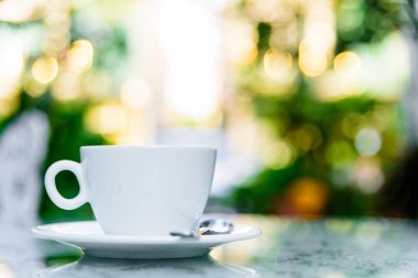 Sabah servis edilecek beyaz bir fincan kahve ve gümüş kaşık. Rahatlatıcı ve sarı ışıklandırma bokeh arka planlı..