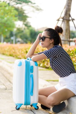 Güzel gezgin Asyalı kadın, büyük beyaz ve mavi bir çantayla sokakta oturuyor. Akşam vakti taksi şoförü bekliyor. Gün batımında..