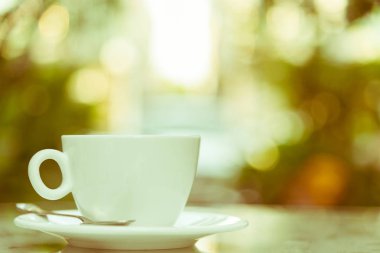 Sabah servis edilecek beyaz bir fincan kahve ve gümüş kaşık. Rahatlatıcı ve sarı ışıklandırma bokeh arka planlı..
