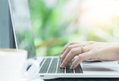 Asyalı kadın elleri ve evlilik yüzüğü laptopta kahve, bilgisayar ve pencerenin dışındaki manzarayla dokunup daktilo ediyor..