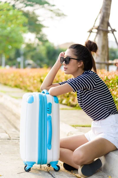 Güzel gezgin Asyalı kadın, büyük beyaz ve mavi bir çantayla sokakta oturuyor. Akşam vakti taksi şoförü bekliyor. Gün batımında..