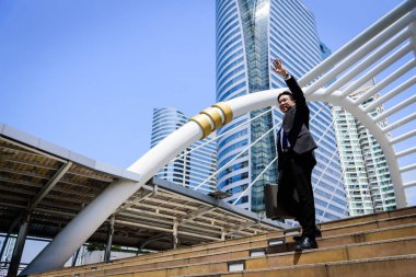 Asyalı işadamının elinde siyah bir çanta var ve merhaba işareti yapıyor..