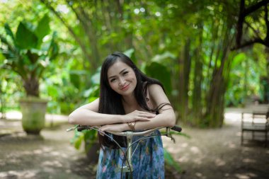 Asyalı güzel bir kız bisiklete biniyor ve mutlu bir şekilde gülüyor. Küçük Ağaç Bahçesi Kafesi, Nakhon Pathom, Tayland..
