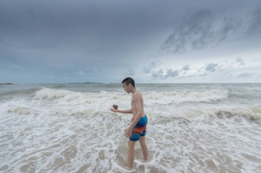 Asyalı bir çocuk kumsalda bulutlu bir gökyüzü ile bir taş tutmalı..