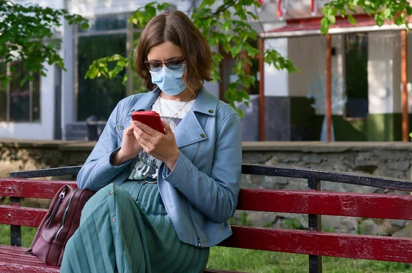 Koruyucu maskeli genç bir kadın akıllı telefon tutuyor. Yeşil parktaki bankta oturan cep telefonlu bir kadın.