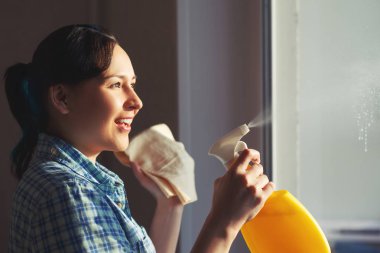 Genç bir kız pencereler yıkar bir süpürge ve mendil bir sünger ile parlak pencerenin dışındaki güneş parlar
