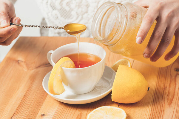 Чашка чая с лимоном на деревянном столе, к которому ложка добавляет мед
