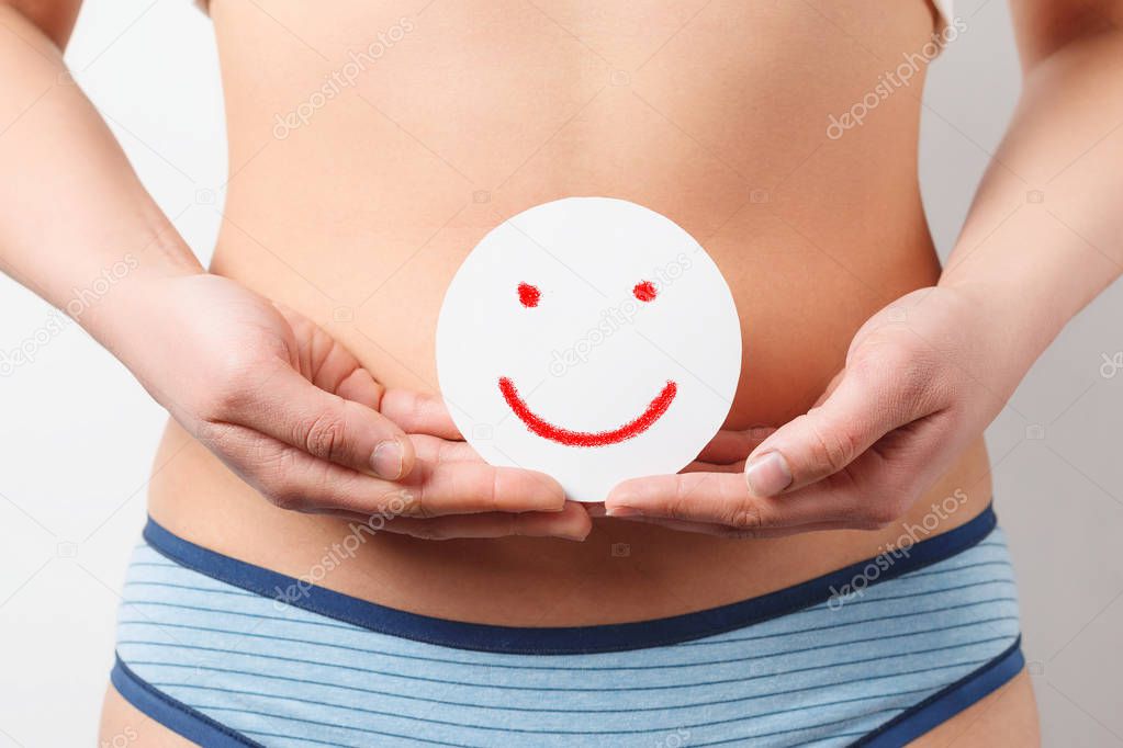 a young woman holds a round card with a smile in her hands at the level of the abdomen. concept of women's health