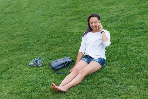 快乐美丽的年轻女子坐在草地上 使用智能手机 — 图库照片