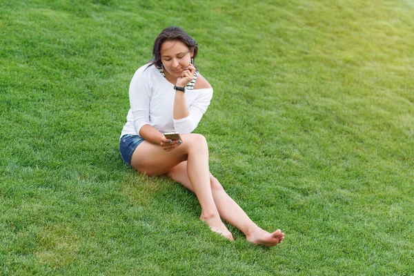 美丽的年轻欧洲女孩坐在公园的草地上 并使用智能手机 — 图库照片