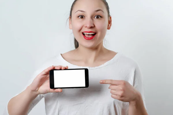 Молодая улыбающаяся женщина держит смартфон горизонтально с белым экраном с местом для копирования и указывает на него пальцем другой руки. на белом фоне — стоковое фото