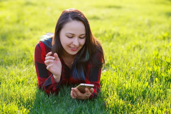 Портрет улыбающейся женщины, лежащей на зеленой траве и использующей смартфон на открытом воздухе — стоковое фото