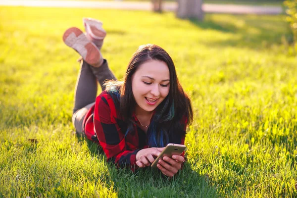 Портрет улыбающейся женщины, лежащей на зеленой траве и использующей смартфон на открытом воздухе — стоковое фото