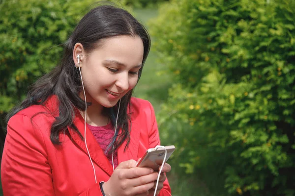 Девушка в красной куртке в парке слушает музыку с наушниками в руках со смартфоном — стоковое фото