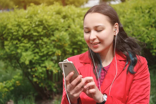 Девушка в красной куртке в парке слушает музыку с наушниками в руках со смартфоном — стоковое фото