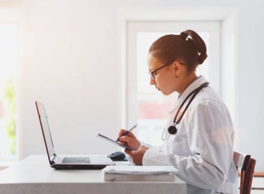 İş yerindeki kadın doktor dizüstü bilgisayar ve not panosuyla çalışıyor.