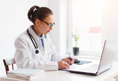 İş yerindeki kadın doktor dizüstü bilgisayar ve not panosuyla çalışıyor.