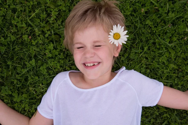 Dítě leží na zeleném trávníku. Blonďatý chlapec se usměje a podívá se do kamery. Blondýnka obklopená květy jetele. Mezera pro text. — Stock fotografie