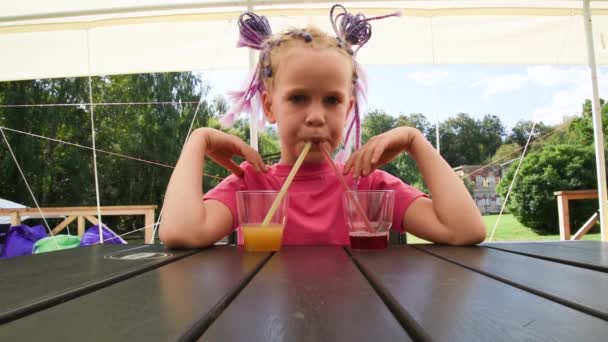 Дівчина з незвичайною красивою зачіскою сидить за столом у літньому кафе. Дитина п'є сік через трубку. Дівчина п'є барвисті напої з трьох окулярів одночасно . — стокове відео