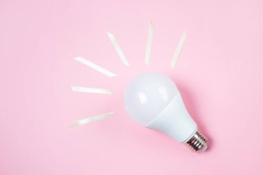 Öndeki lamba pastel pembe bir arka planda yatıyor. Yakındaki saten sarı kurdele parçaları lambadaki ışık ışınlarını sembolize eder. Fikir konsepti. Minimalizm, üst görünüm, düz uzanma, metin için yer.