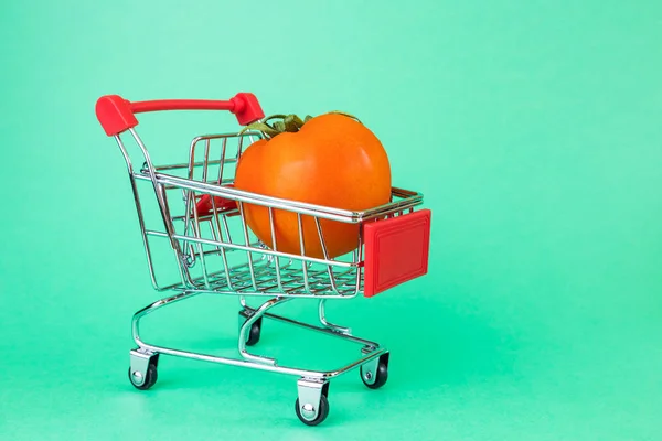A small copy of a supermarket basket on a green background. Inside is a ripe tomato. The concept of shopping, grocery stores, discounts, promotions.