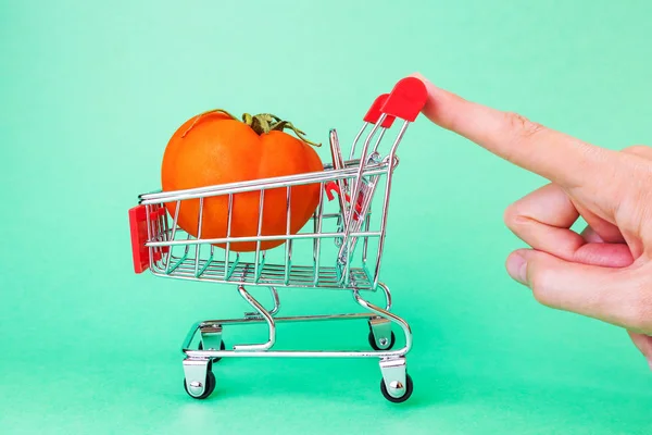 A small copy of a supermarket basket on a green background. Inside is a ripe tomato. The concept of shopping, grocery stores, discounts, promotions.