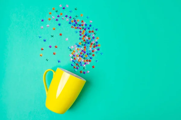 A yellow mug on a green background, near - rhinestones in the shape of stars. The concept of a holiday, party, new year. Top view, flat lay. Minimalism. Copy space.