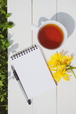 Yaz aylarında, bir fincan çay, boş sayfalı bir not defteri, bir kalem ve beyaz ahşap arka planda üç sarı papatya. Yanında taze otlar var. Parlak güneş ışığı. Boşluk, minimalizm.