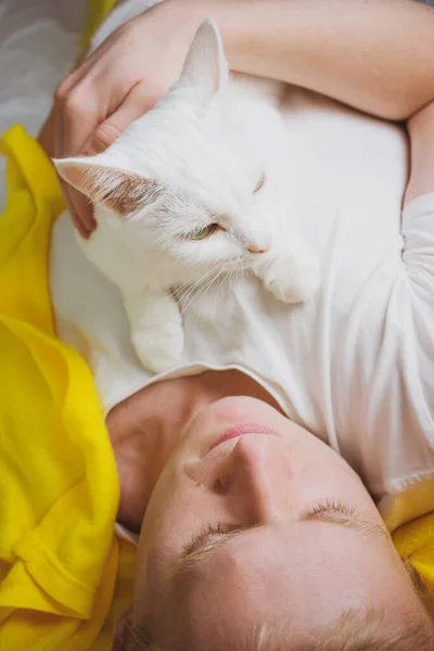 A young blond man is lying on a bed with a white shorthair cat. The concept of pets, weekends, laziness, morning. Photo in bright white and yellow tones.