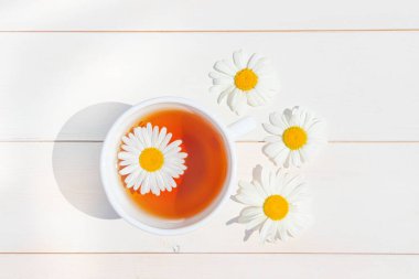 Hafif ahşap bir yüzey üzerinde küçük beyaz bir fincanda papatya çayı. Fincanın etrafında dağınık çiçekler var. Sağlık ve bağışıklık için doğal vitamin içecekleri konsepti. Üst manzara, düz uzanma.