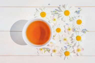 Hafif ahşap bir yüzey üzerinde küçük beyaz bir fincanda papatya çayı. Fincanın etrafında dağınık çiçekler var. Sağlık ve bağışıklık için doğal vitamin içecekleri konsepti. Üst manzara, düz uzanma