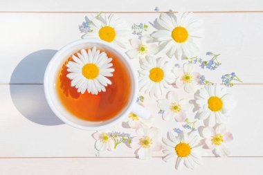 Hafif ahşap bir yüzey üzerinde küçük beyaz bir fincanda papatya çayı. Fincanın etrafında dağınık çiçekler var. Sağlık ve bağışıklık için doğal vitamin içecekleri konsepti. Üst manzara, düz uzanma