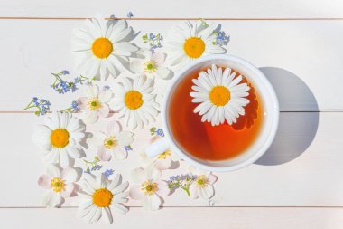 Hafif ahşap bir yüzey üzerinde küçük beyaz bir fincanda papatya çayı. Fincanın etrafında dağınık çiçekler var. Sağlık ve bağışıklık için doğal vitamin içecekleri konsepti. Üst manzara, düz uzanma.