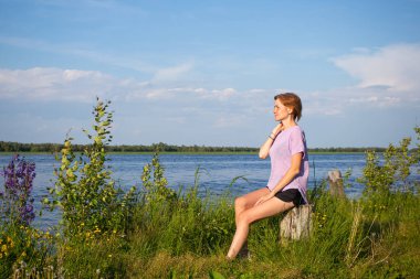 Yalnız yürüme kavramı. Mor tişörtlü ve şortlu kısa saçlı genç kız nehir kıyısında oturuyor. Yaz güneşli bir akşam. Kopyalama. Hoş, şirin kırsal alan.