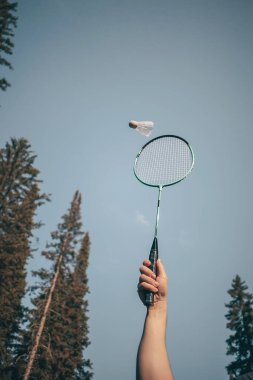 Bir el badminton raketini tutar, bir mekik horozu yakınlarda uçar. Amatör badminton oyunu konsepti, açık hava aktiviteleri. Kopyalama alanı.