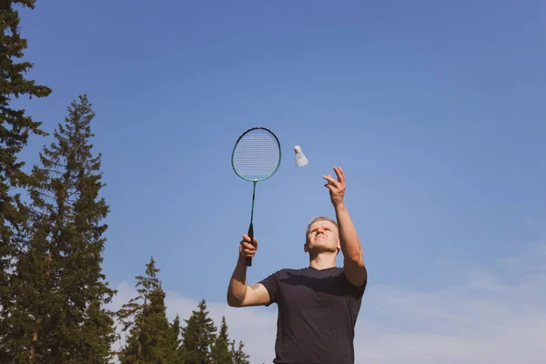 Young Caucasian man plays badminton on a background of blue sky and forest. The concept of an amateur game of badminton, outdoor activities. Copyspace, bottom view.