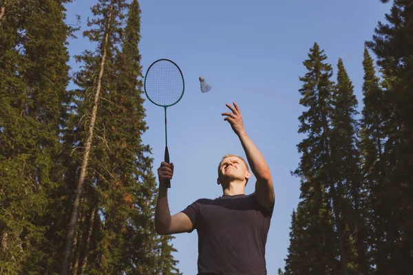 Young Caucasian man plays badminton on a background of blue sky and forest. The concept of an amateur game of badminton, outdoor activities. Copyspace, bottom view.