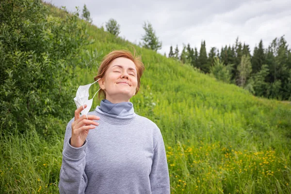 Concept of quarantine ending, walks alone. Young women are taking off a disposable medical mask in a park on a background of a hill with grass and trees.