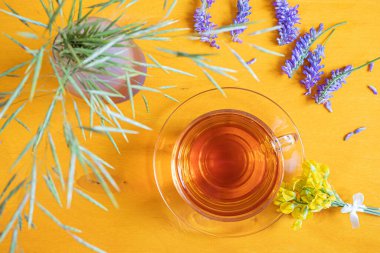 Çayı şeffaf bir fincanda çay, çay tabağı, tarla bitkileri, sarı ahşap arka planda sarı ve mor çiçekler. Yukarıdan bak. Tatlı yaz yatağı.