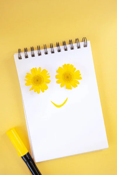 Notebook on a spiral. Creative concept of a smiley face. A smile is drawn on the page with a yellow marker. Two yellow daisies as eyes.