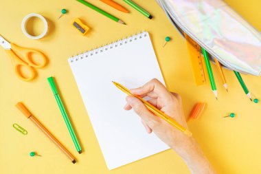 Okula dönüş kavramı. Not defteri, suluboya, kalemler, kalemler, keskinleştirici, makas, cetvel, sarı arkaplanda ataçlar. Elin kalem tutsun. Turuncu ve yeşil renkler. Kopyalama alanı.