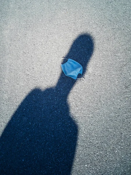 The used disposable medical mask is lying on the pavement. The shadow of a departing person. The concept of covid, coronavirus, garbage, urban pollution.